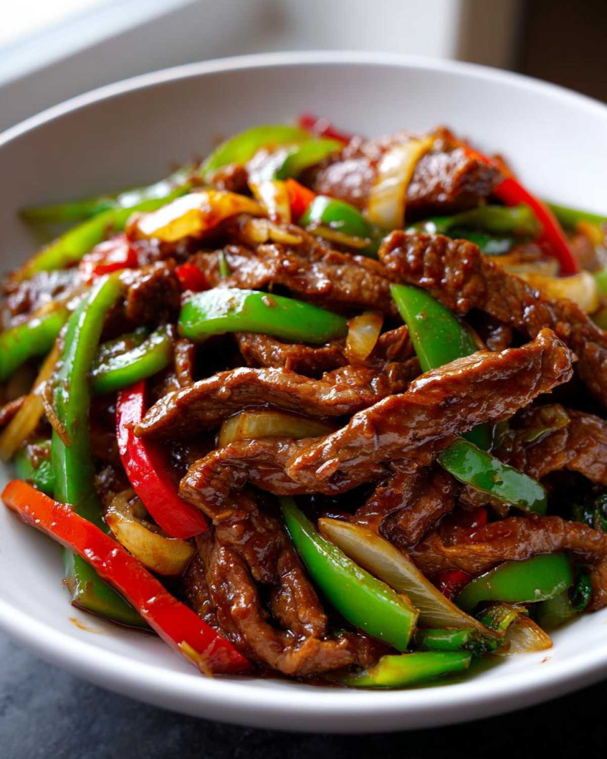 Close-up of tender strips of beef coated in savory sauce alongside bright green and red peppers in a bowl of pepper steak.