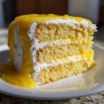Close-up of a moist slice from an amazing lemon cake recipe, layered with white frosting and drizzled with bright yellow lemon glaze.