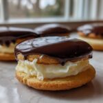 Close-up of a boston cream pie cookie sandwich filled with cream and topped with dripping dark chocolate glaze.