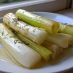 Close-up of tender braised leeks seasoned with rosemary, sitting in a light sauce on a white plate.