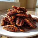 A close-up, sunlit pile of glossy, dark brown candied pecans recipe served on a small white plate.