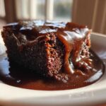 A close-up of a rich, dark slice of chocolate pudding cake covered in glossy chocolate sauce.