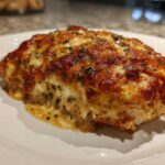 A close-up of a baked caesar chicken breast covered in melted, browned cheese and herbs.