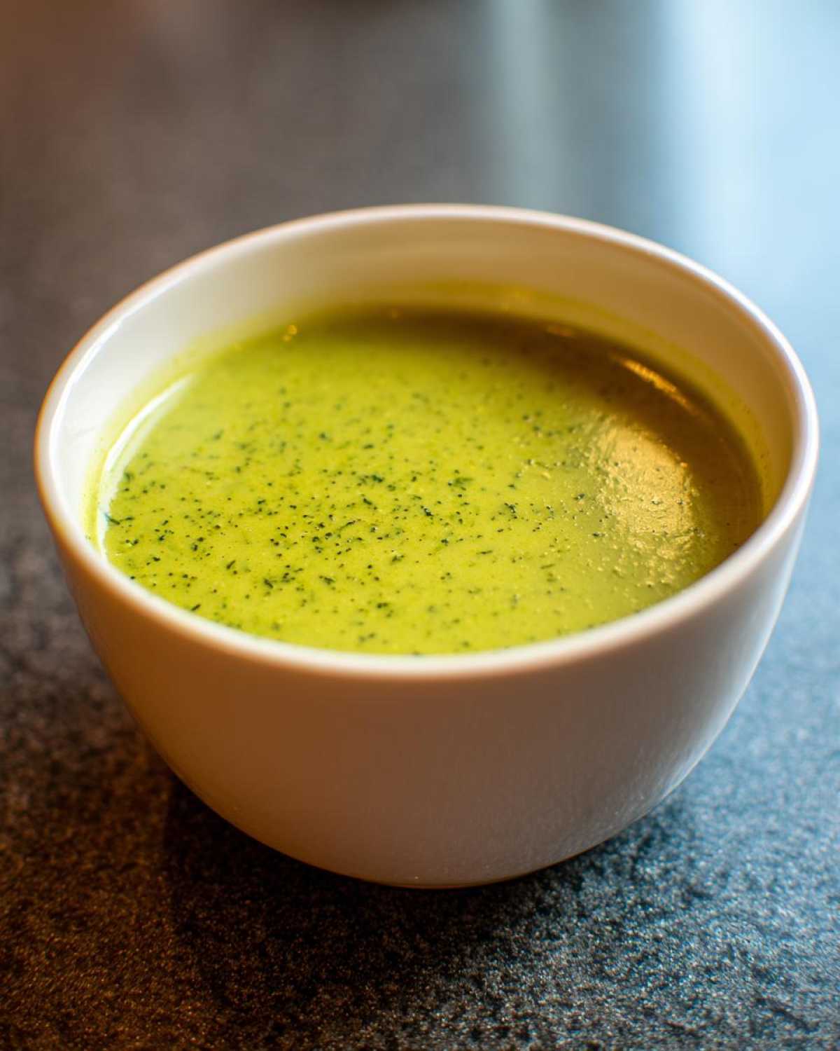 Close-up of a bowl of vibrant green, creamy zucchini soup, garnished with dark herbs.
