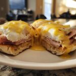 Close-up of two halves of eggs benedict showing poached egg, ham, and rich hollandaise sauce dripping onto a white plate.