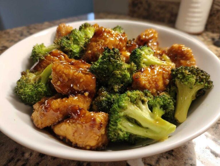 A white bowl filled with saucy, golden pieces of chicken and bright green broccoli florets, featuring the main dish: chicken and broccoli.