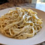 A close-up of fettuccine pasta generously coated in creamy homemade alfredo sauce and topped with cracked black pepper.