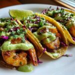 Three perfectly grilled shrimp tacos topped with purple cabbage, cilantro, and a creamy green sauce, served with a lime wedge.
