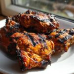 Close-up of beautifully charred and glazed Jamaican jerk chicken pieces piled on a white plate near a window.
