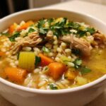 Close-up of a white bowl filled with hot penicillin soup featuring shredded chicken, carrots, celery, and barley, topped with fresh parsley.