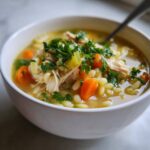 Close-up of a white bowl filled with penicillin soup featuring shredded chicken, carrots, orzo pasta, and fresh parsley garnish.