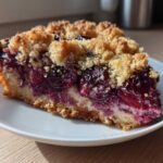 A close-up of a moist slice of plum cake featuring dark, juicy fruit filling and a golden, crunchy streusel topping.