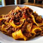 Close-up of wide, yellow pappardelle pasta generously topped with rich, meaty bolognese sauce in a white bowl.