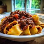 A close-up of wide pappardelle pasta generously topped with rich, meaty bolognese sauce in a white bowl.