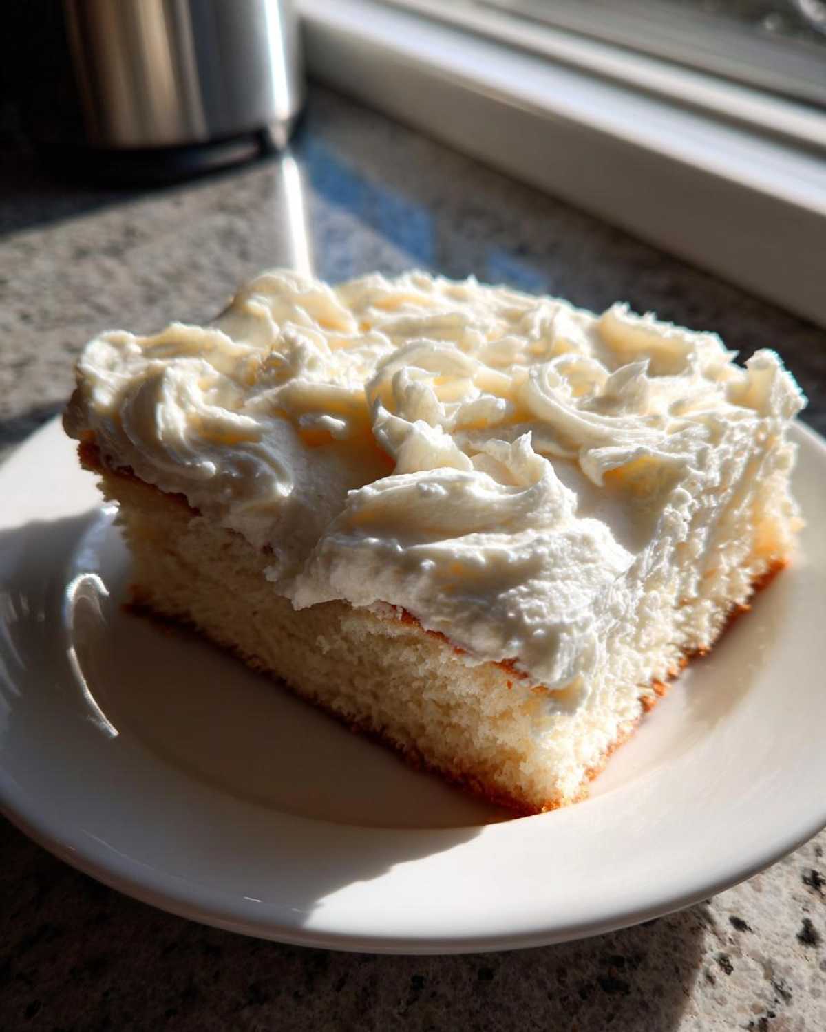 A square slice of white cake generously topped with swirls of fluffy vanilla buttercream frosting, sitting on a white plate.