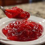 A spoonful of glistening, bright red strawberry freezer jam being lifted above a small white bowl of the jam.