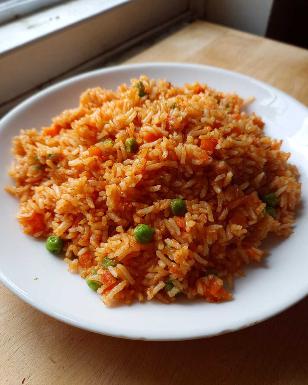 A close-up of a serving of vibrant orange spanish rice mixed with bright green peas and diced carrots on a white plate.