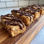 A row of homemade, oat-based protein bars drizzled with melted chocolate, displayed on a wooden cutting board.