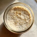 Overhead view of an active sourdough starter with many bubbles and specks of flour in a glass jar.