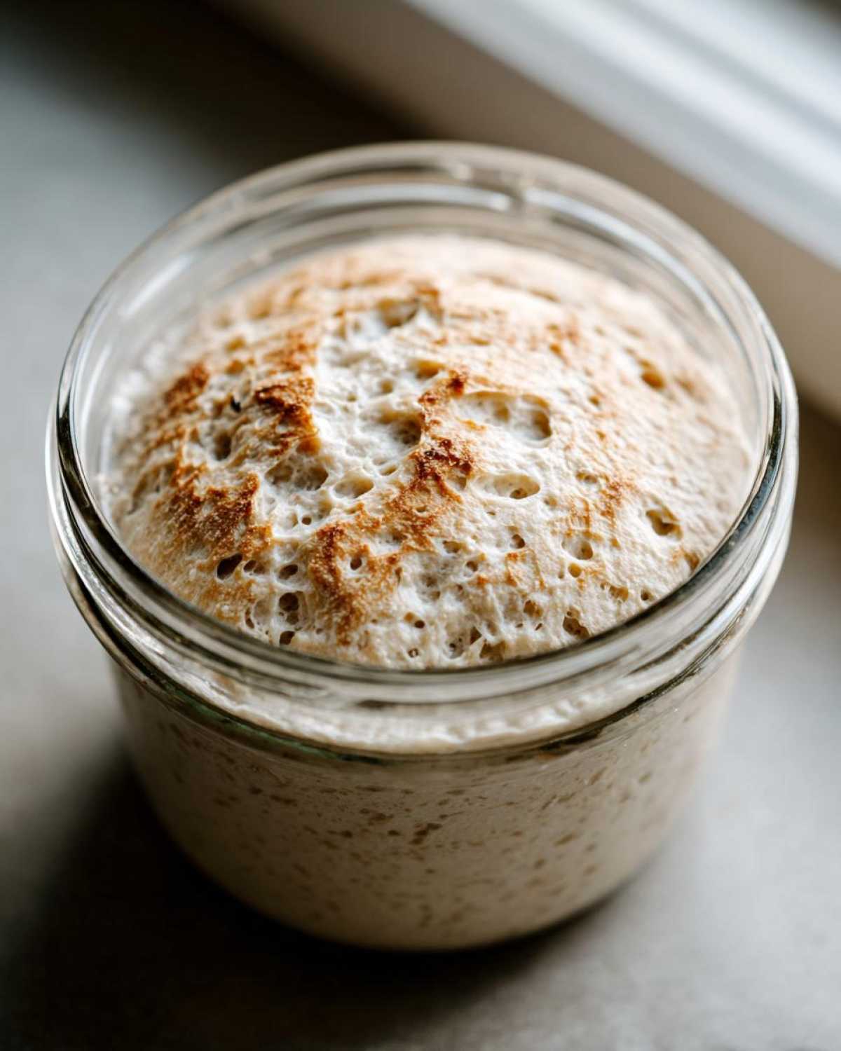 Close-up of a very active sourdough starter, risen high in a glass jar with a bubbly, golden-brown top.
