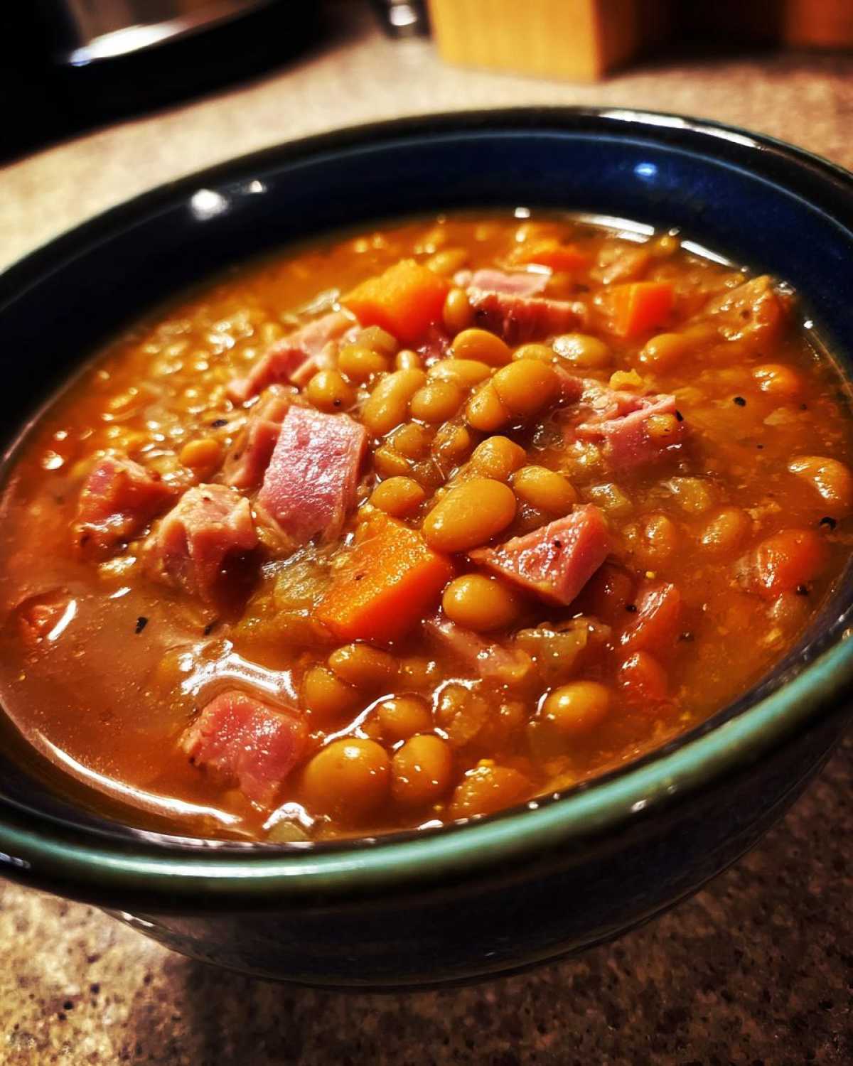 Close-up of a dark blue bowl filled with rich, thick 15 bean soup, featuring visible ham chunks and carrots.