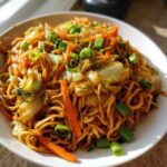 A close-up, appetizing shot of a white bowl filled with savory fried noodles, cabbage, carrots, and green onions.