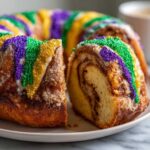 A slice cut from a homemade king cake, showing the cinnamon swirl interior and purple, green, and gold sprinkles.