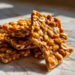Close-up of shiny, golden-brown pieces of peanut brittle recipe stacked on a marble surface.
