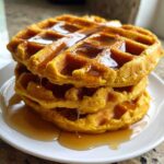 A close-up of three stacked, golden-orange pumpkin waffles drizzled generously with maple syrup.