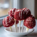 A cluster of vibrant red velvet cake pops coated in crumbs and drizzled with white chocolate, standing in a small white bowl.