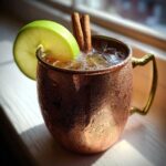 A frosty copper mug filled with an apple cider moscow mule, garnished with a green apple slice and cinnamon sticks.