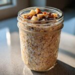 Close-up of apple cinnamon overnight oats soaking in milk inside a glass jar, topped with diced apples and cinnamon.