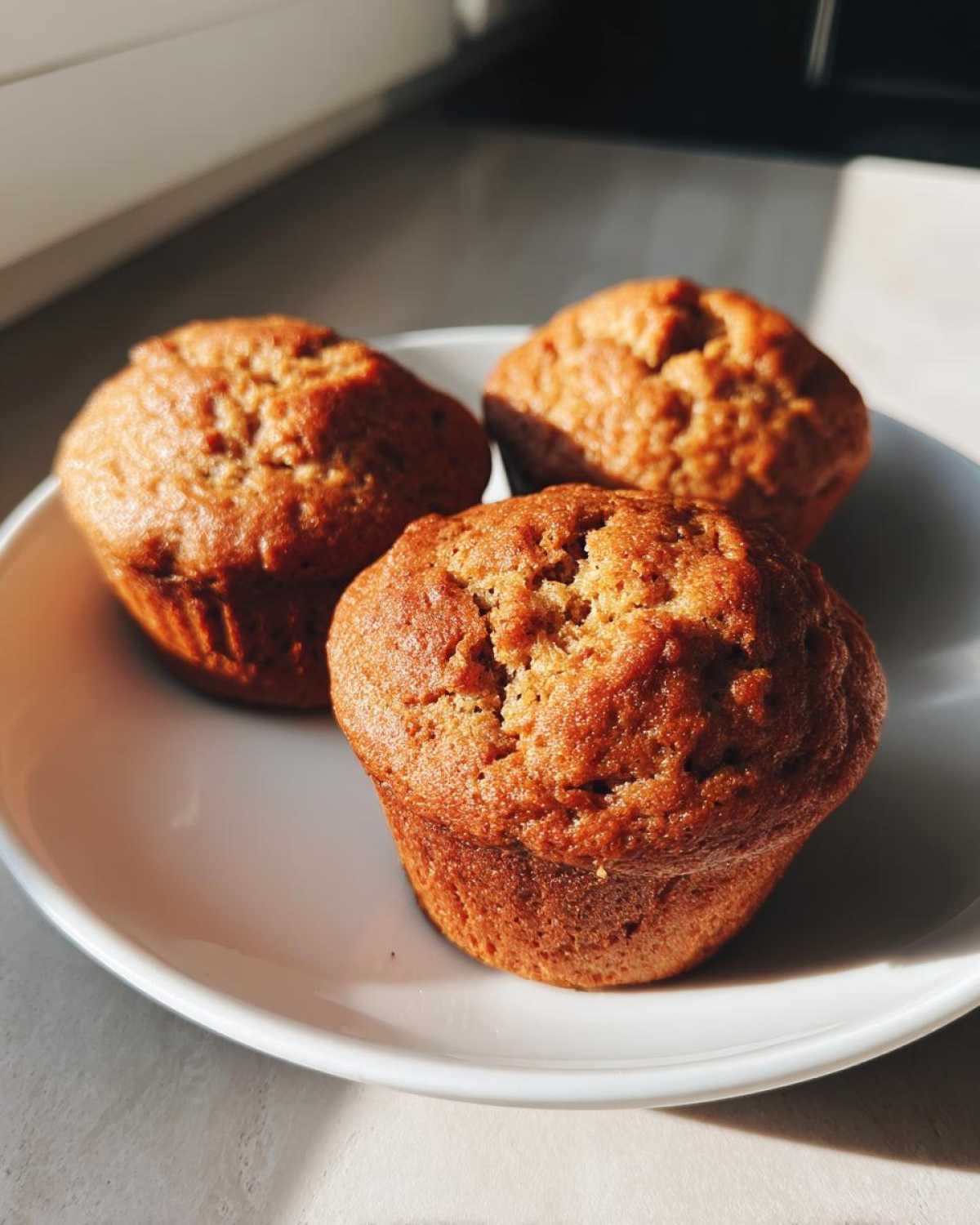 Three freshly baked banana protein muffins resting on a white plate, highlighted by natural sunlight.