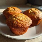 Three freshly baked banana protein muffins sitting on a white plate, illuminated by natural sunlight.