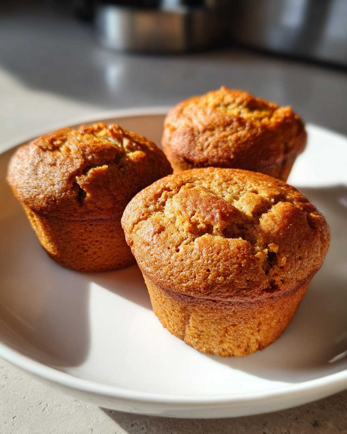 Three freshly baked, golden brown banana protein muffins sitting on a white plate in bright sunlight.