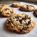 Close-up of a freshly baked chocolate chip cookie topped with flaky sea salt, resting on parchment paper.