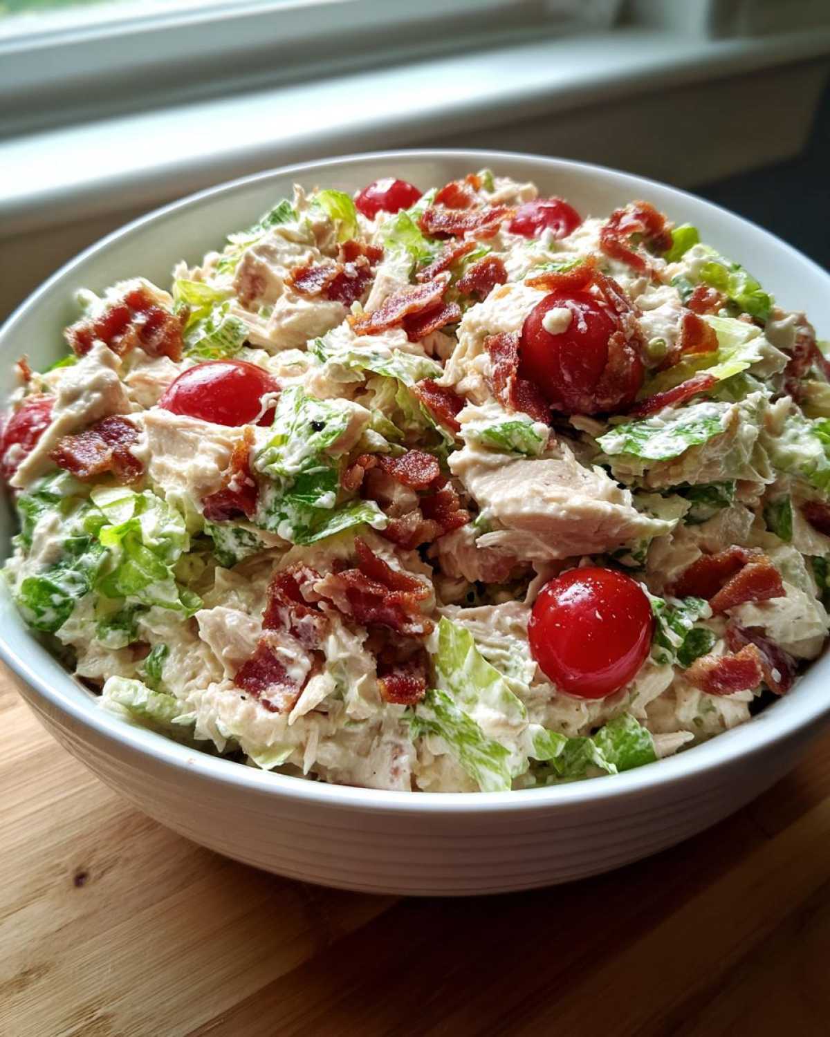 A close-up of a white bowl filled with creamy blt chicken salad, topped with crispy bacon bits and cherry tomatoes.