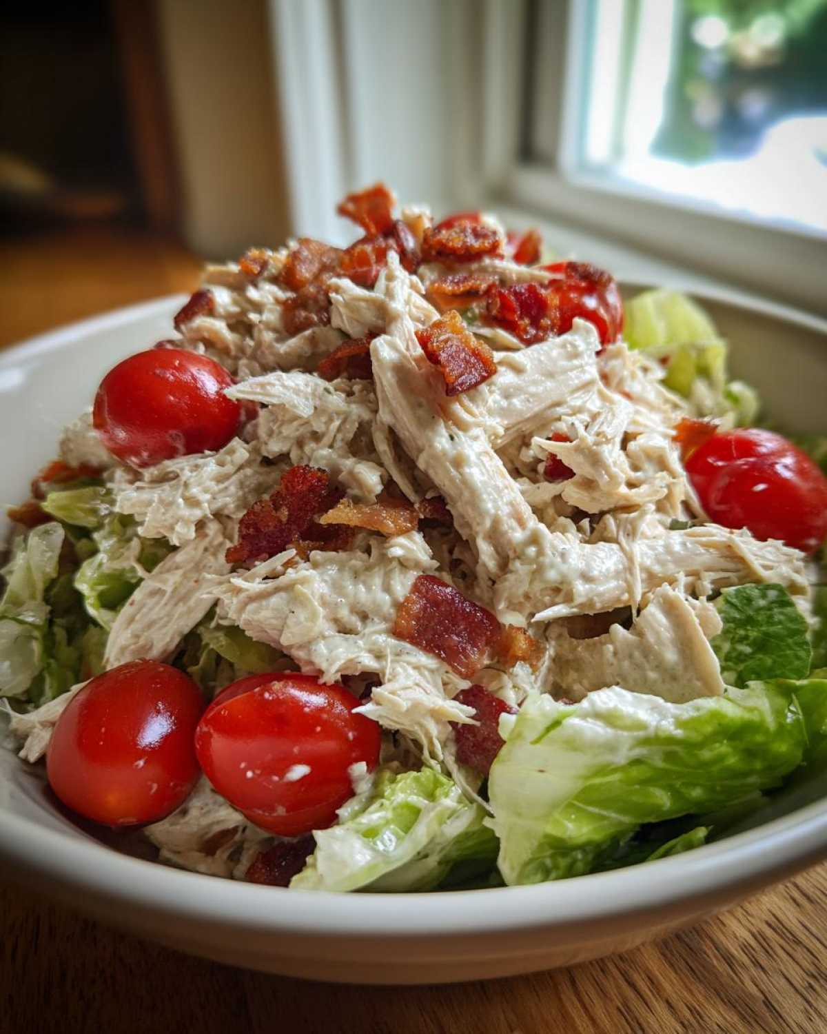 A bowl filled with creamy blt chicken salad, shredded chicken, crispy bacon bits, and cherry tomatoes over lettuce.