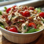 A close-up view of a creamy blt chicken salad topped with crispy bacon bits and bright red cherry tomatoes, served over lettuce.