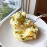 A fork lifting a bite of creamy Boursin mashed potatoes mixed with fresh green herbs.