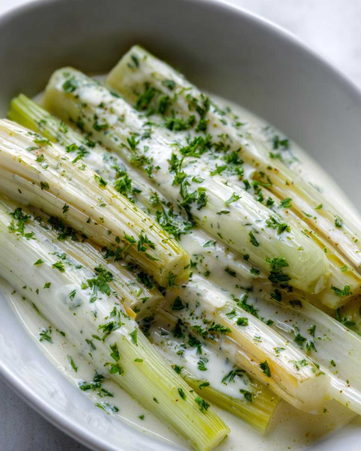 Close-up of tender braised leeks covered in a creamy white sauce and sprinkled with fresh parsley.