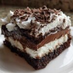 A close-up of a rich, layered brownie refrigerator cake slice topped with whipped cream and chocolate shavings.