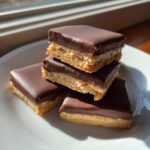 Four square pieces of buckeye graham crackers stacked on a white plate, featuring a graham base, peanut butter filling, and thick chocolate coating.