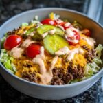 A close-up view of a delicious cheeseburger bowl featuring ground beef, melted cheddar, pickles, tomatoes, and special sauce over lettuce.