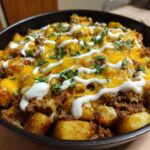 Close-up of Cheesy Taco Potatoes topped with melted cheddar, sour cream drizzle, and cilantro in a black skillet.