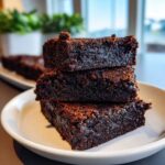 A stack of three chewy gingerbread brownies on a white plate, showing rich dark texture.