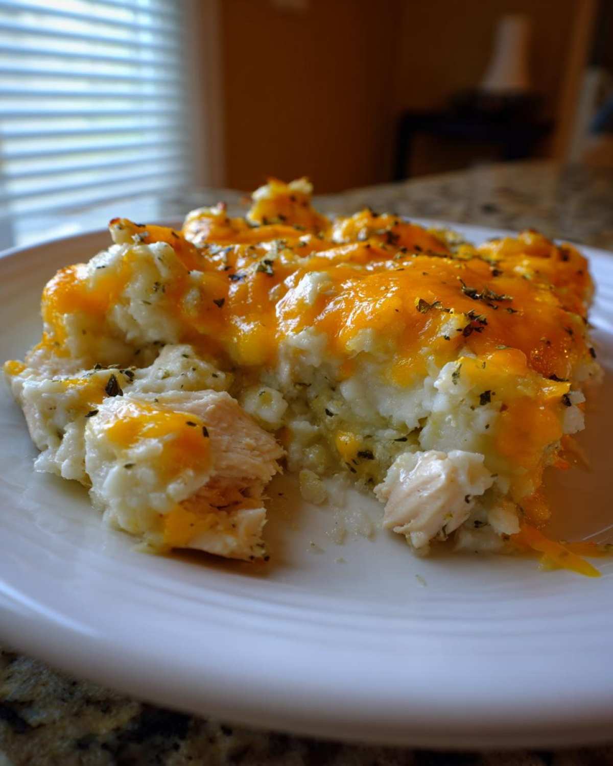A close-up serving of chicken cauliflower rice casserole topped with melted cheddar cheese and herbs on a white plate.