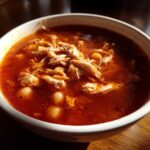 Close-up of a white bowl filled with steaming, red broth chicken pozole, featuring shredded chicken and hominy kernels.