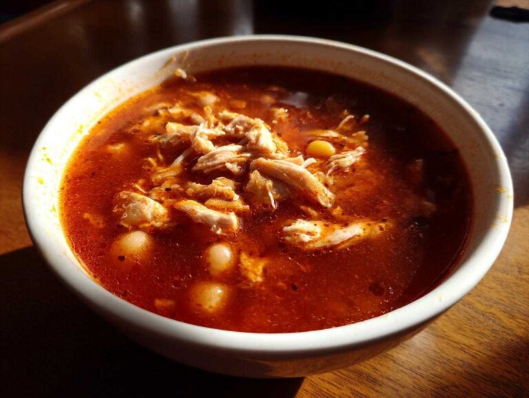 Close-up of a white bowl filled with steaming, red broth chicken pozole, featuring shredded chicken and hominy kernels.