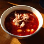A close-up of a white bowl filled with rich, red broth chicken pozole, topped with shredded chicken and hominy.